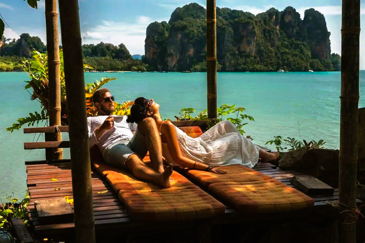Couple relaxing on wooden loungers by turquoise water, cliffs in background.