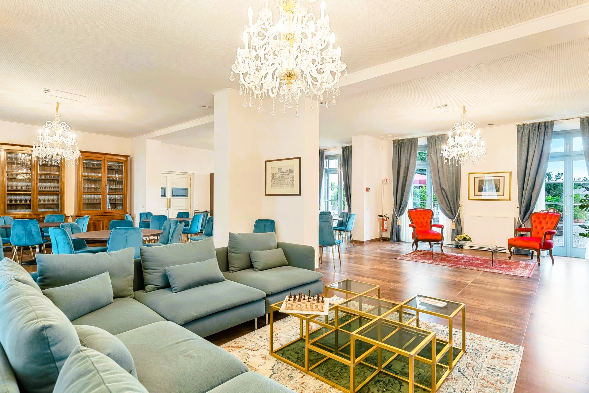 Elegant lounge with blue sofa, chairs, chandeliers, wood floor, and garden view.