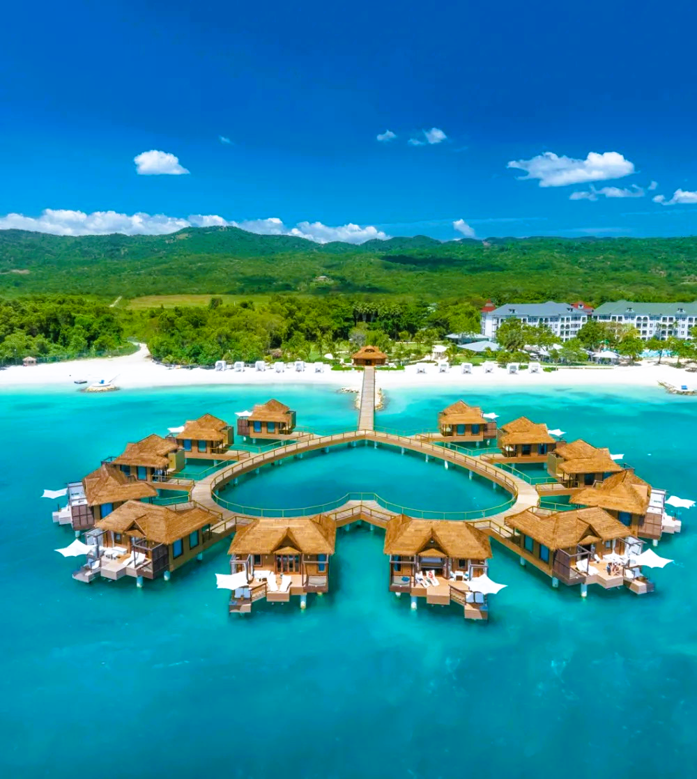 Overwater bungalows in a heart shape in turquoise water, Jamaica. Lush green mountains and white sand beach in background.