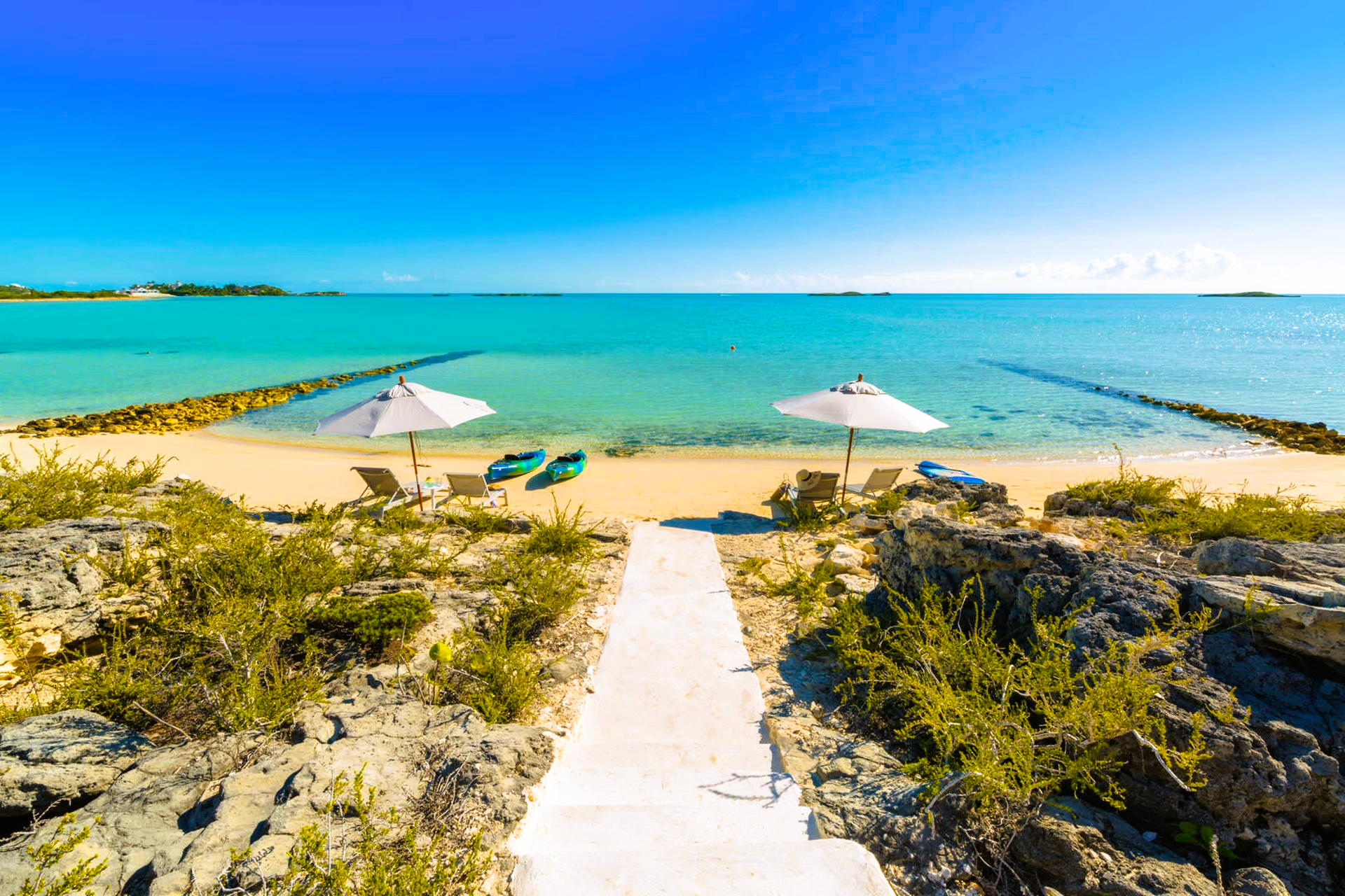 A white path leading to a beach with umbrellas and chairs.
