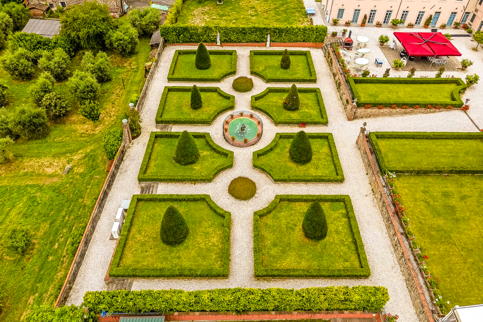 Aerial view of a formal garden with trimmed hedges, symmetrical layout, and a small fountain in the center.