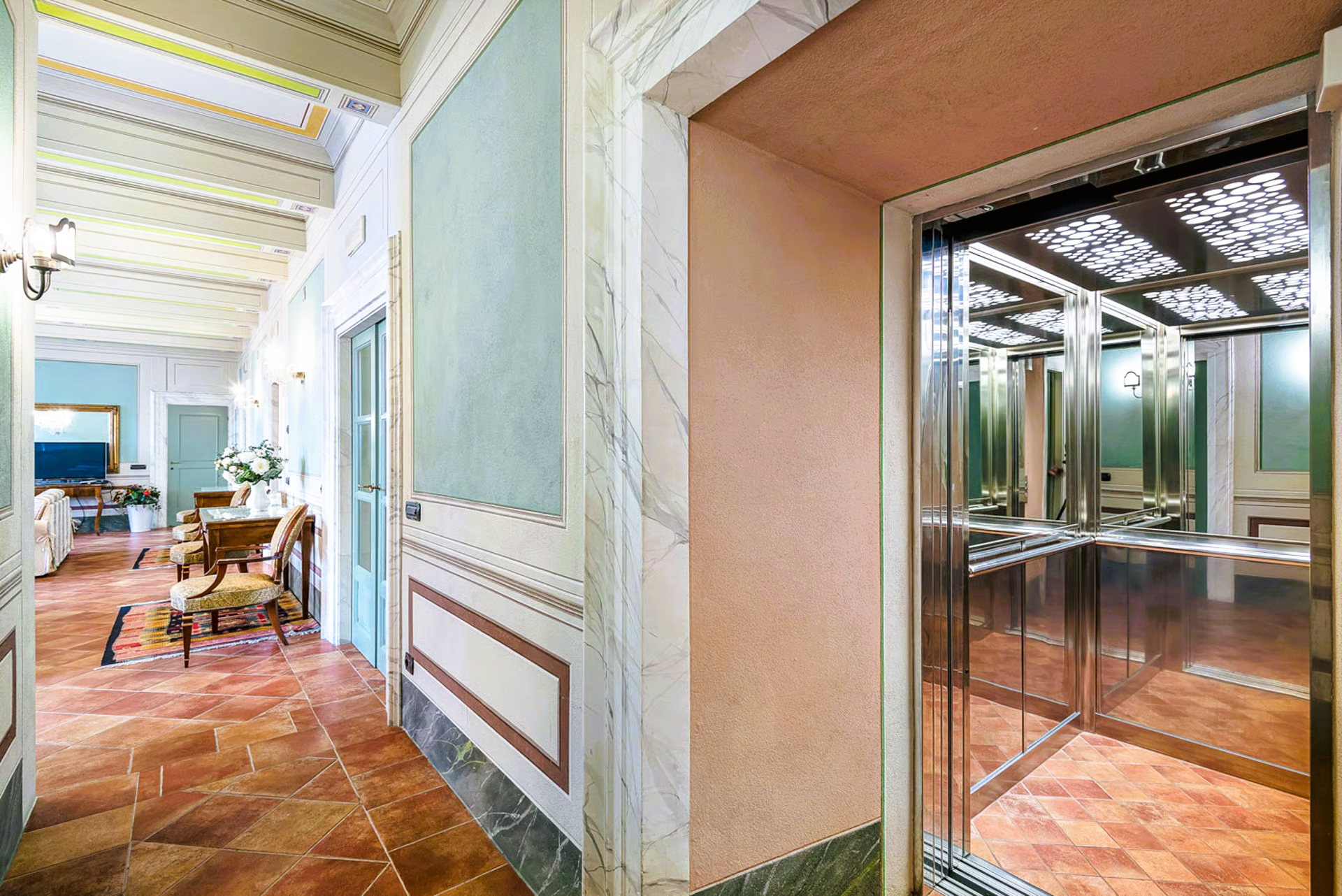 Hallway with terracotta tile floor, elevator with glass walls, and pastel-painted walls.