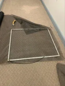 A window screen frame with new mesh, utility knife, and roll of screen material on a carpeted floor.