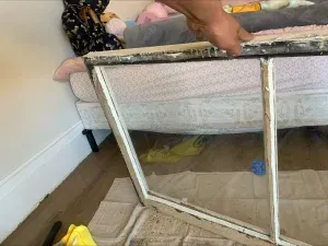 Person holding a white window frame with glass, next to a bed and floor.