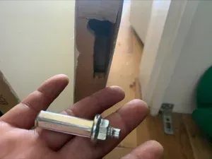 Hand holding a metal bolt, likely from a door frame, near a partially damaged door and wooden floor.