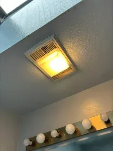 Ceiling light fixture with a bright, square light and vent. Below it, a mirror with multiple lightbulbs.