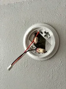 White ceiling-mounted smoke detector base with wires exposed, mounted on a white textured wall.