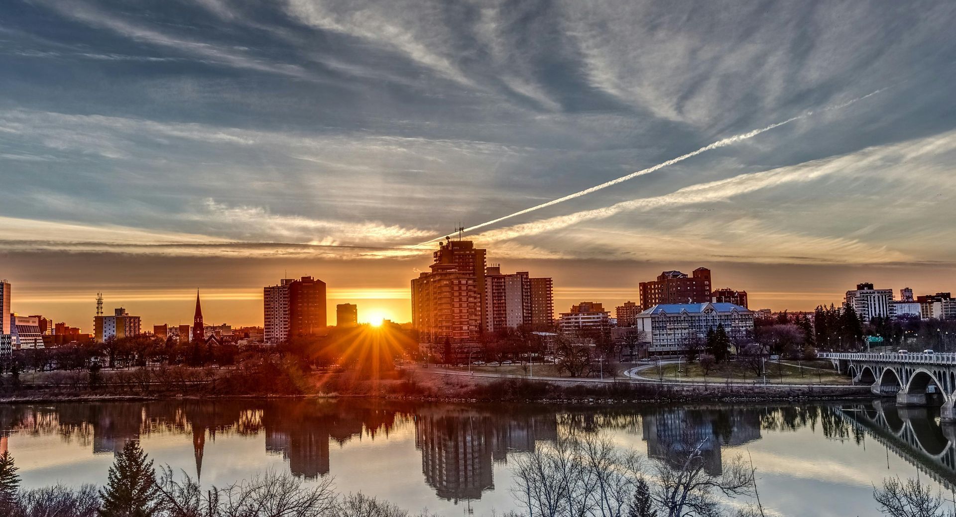 A city skyline is reflected in a lake at sunset.
