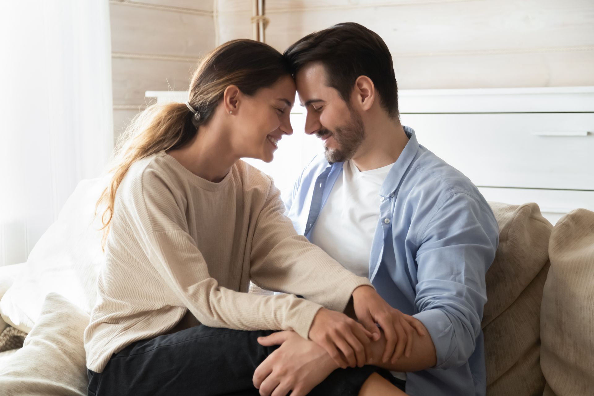 Couple touching foreheads, smiling, and holding hands on a sofa.
