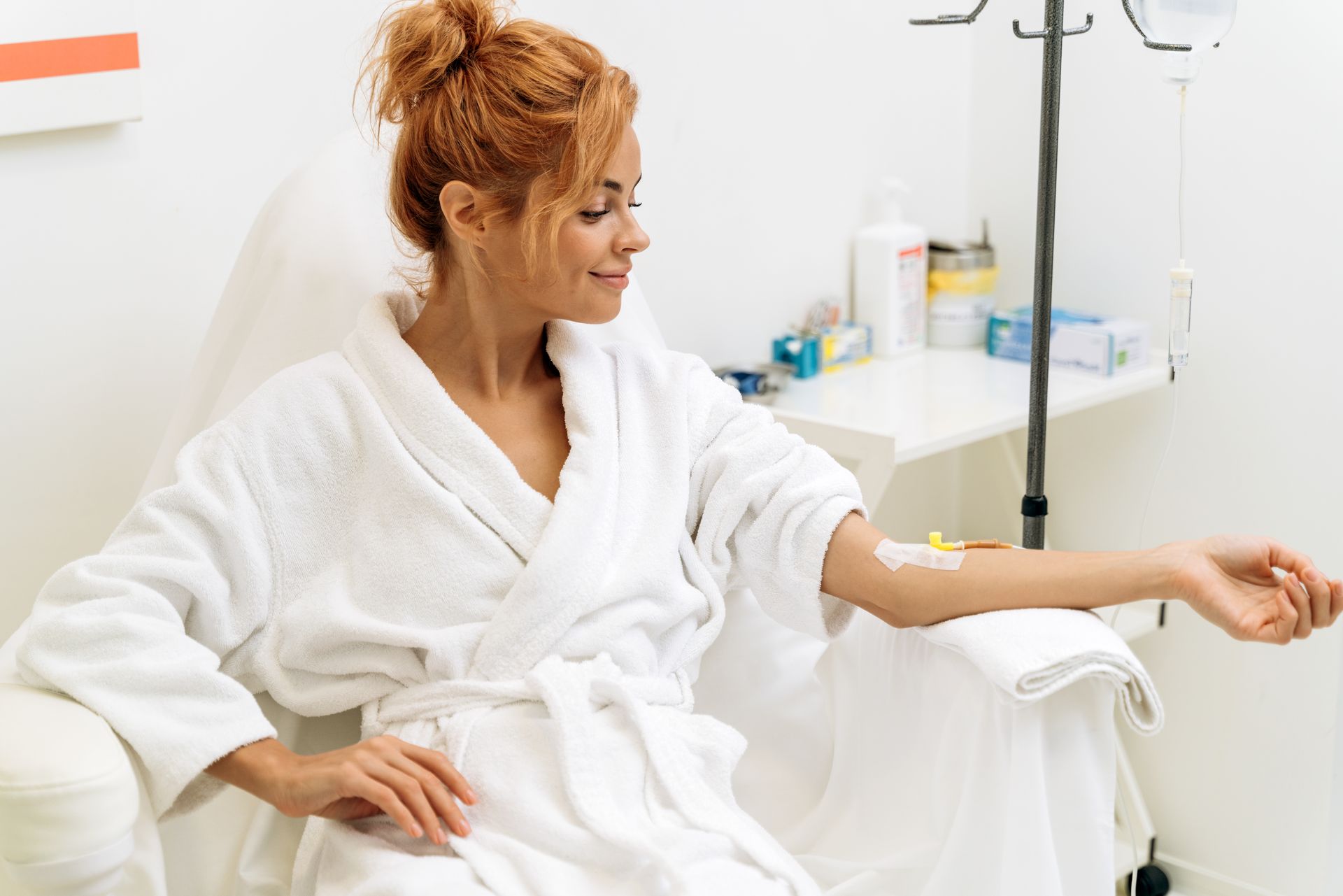 Woman in robe receiving IV drip in a medical setting, smiling.