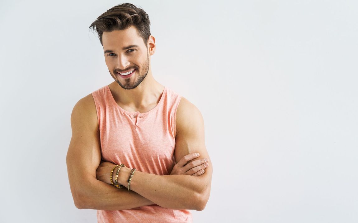 A man in a pink tank top is standing with his arms crossed and smiling.