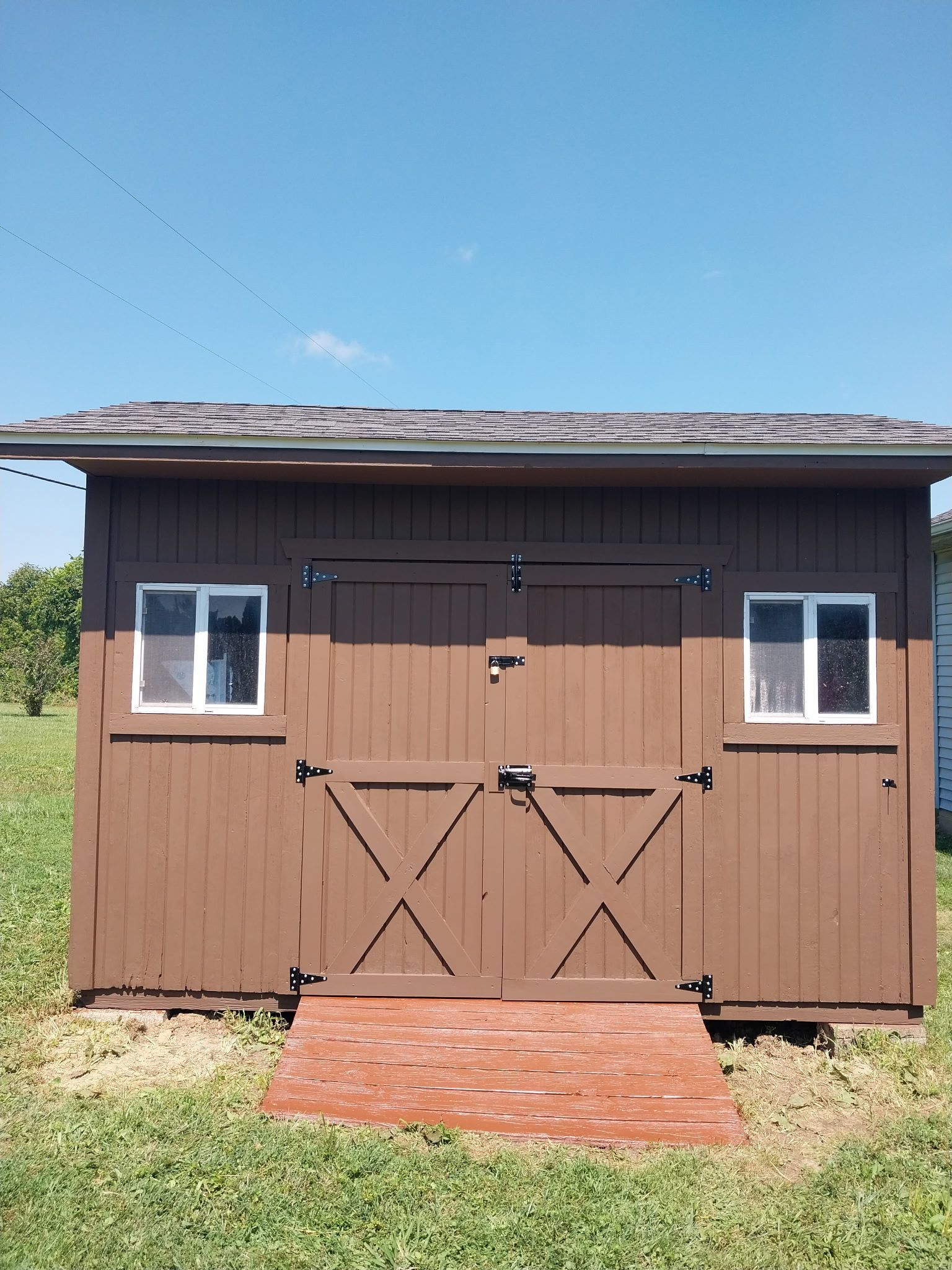 Brown shed with double doors, two windows, and a brick ramp on a grassy field.