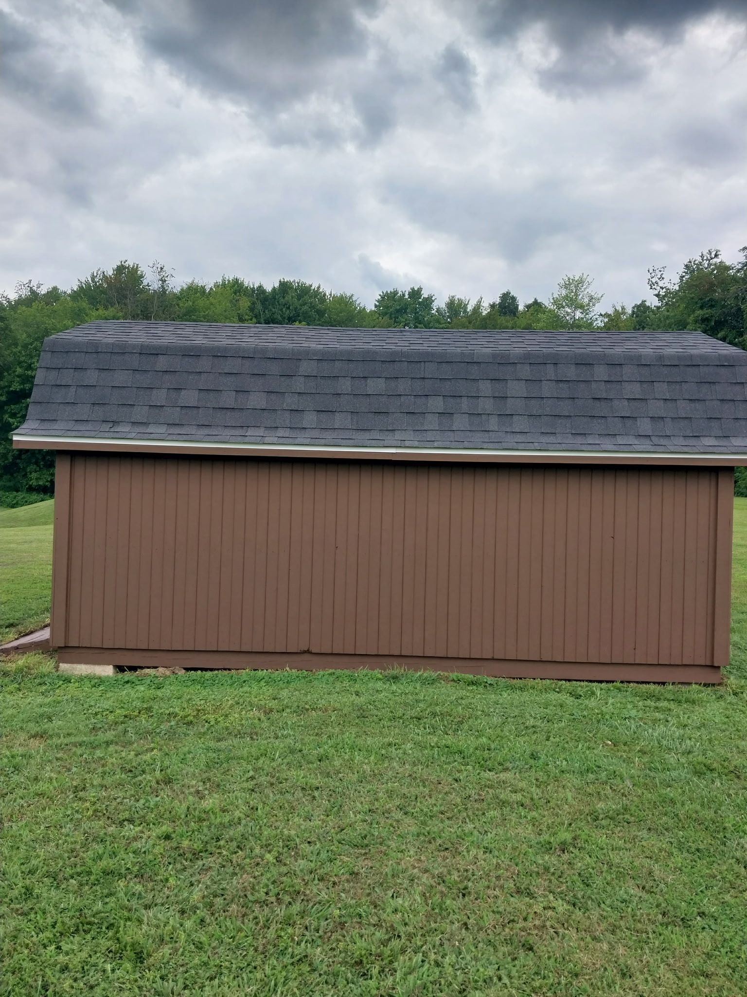 Brown shed with dark gray roof in a grassy field under a cloudy sky.
