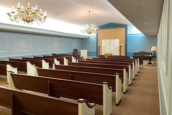 Interior of a chapel with rows of wooden pews, a podium, and chandeliers. Light blue walls.