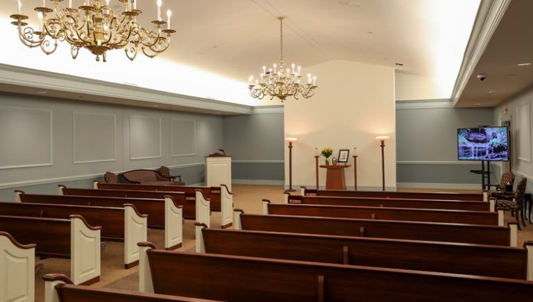 Funeral chapel with rows of pews, chandeliers, and a screen displaying a photo.