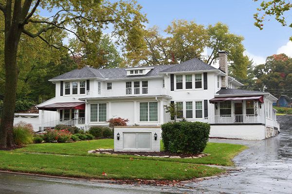 White two-story building with black shutters, a lawn, and a driveway. Red awnings over windows. Trees in background.