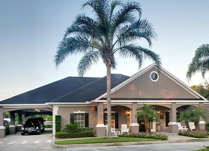 A gray building with a black roof, porch, and a palm tree in front. A hearse is parked under a carport.
