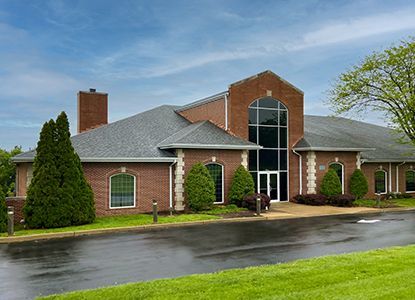 Red brick building with a gray roof, large arched window above double doors. Green bushes and grass surround it.