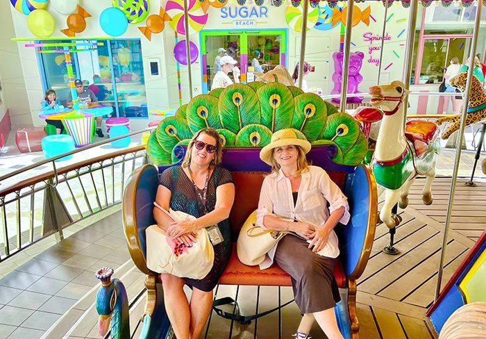 Two women seated on a peacock-themed carousel carriage, smiling. Candy store backdrop.
