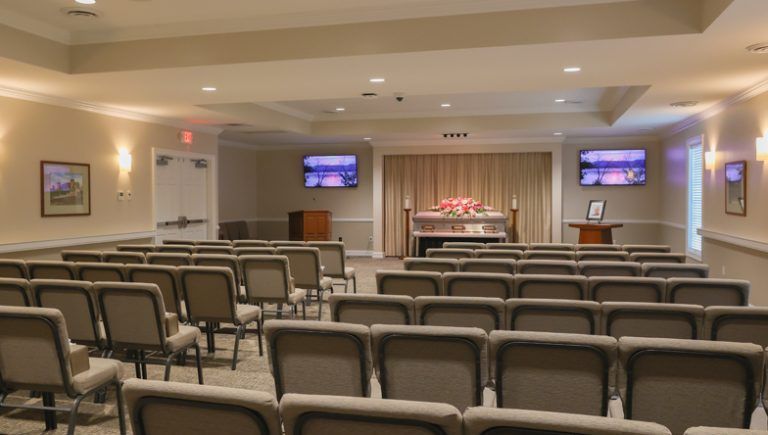 Funeral home chapel with rows of chairs facing a raised platform. Beige walls, soft lighting, and muted colors.