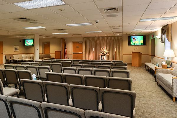 Interior of a funeral home chapel with green chairs, tan walls, and floral arrangements.