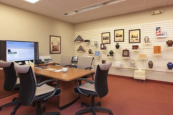 An office with a table and chairs, large screen, and display of urns, flags, and framed items. Red carpet.