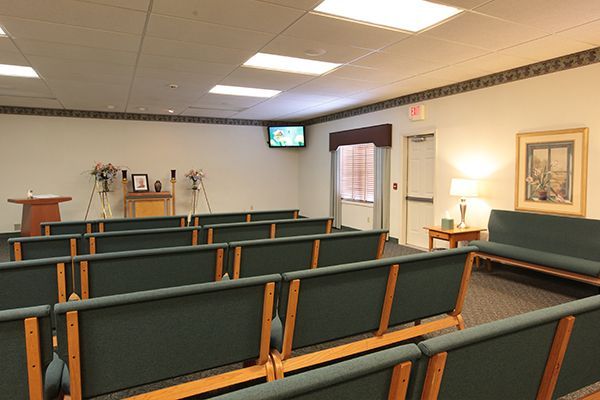 Funeral home chapel: Rows of green cushioned benches face a podium with flowers. A small TV is mounted.