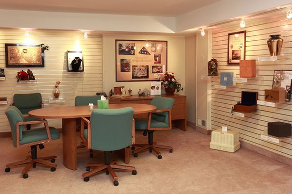 A funeral home consultation room. Beige walls, green chairs, display of urns, photos.