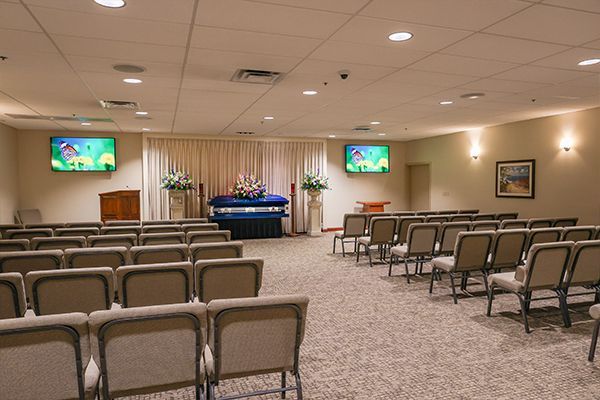 Interior of a funeral home chapel with rows of blue chairs, a floral display, and monitors.