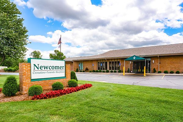 Newcomer Funeral Home in fall setting; brick sign, building, lawn, colorful trees, American flag.
