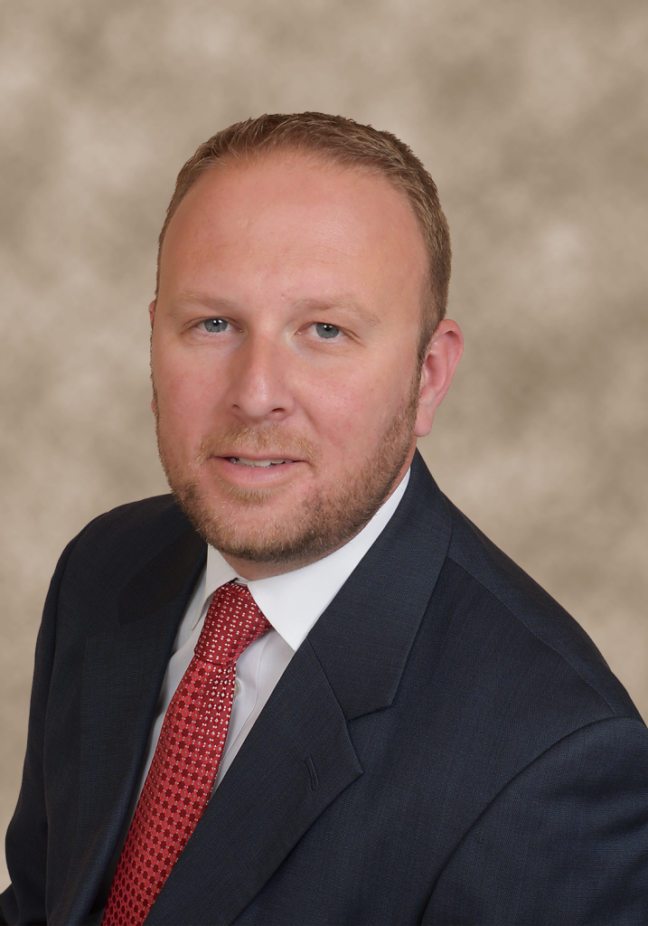 Man in suit, red tie, and light brown hair, smiling at the camera against a neutral background.