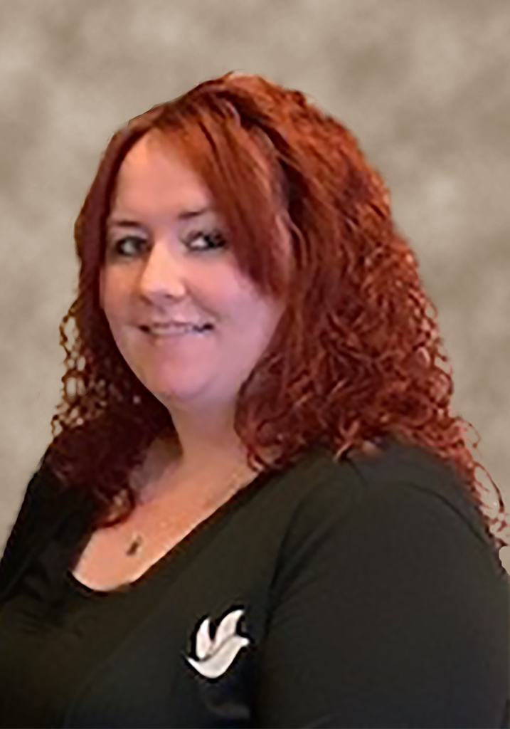 Woman with curly red hair, smiling, wearing a black top with a dove emblem, neutral background.