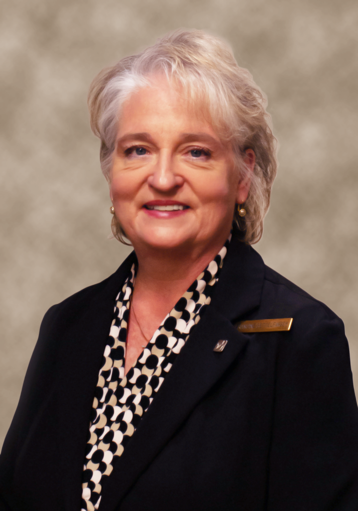 Smiling older woman with gray hair, wearing a black blazer over a black and white patterned blouse.
