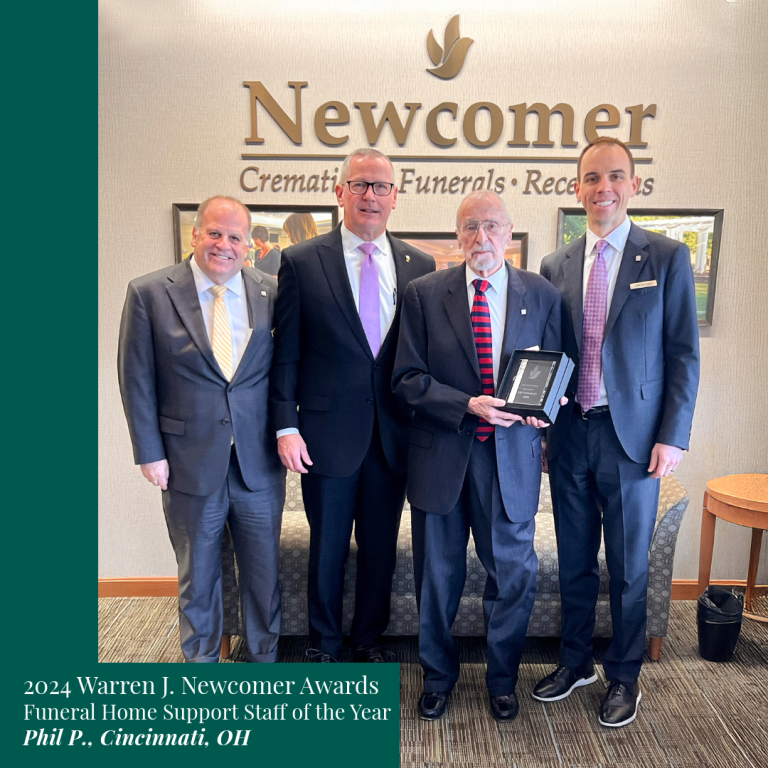 Four men posing for a photo at Newcomer. One holds an award. Text reads