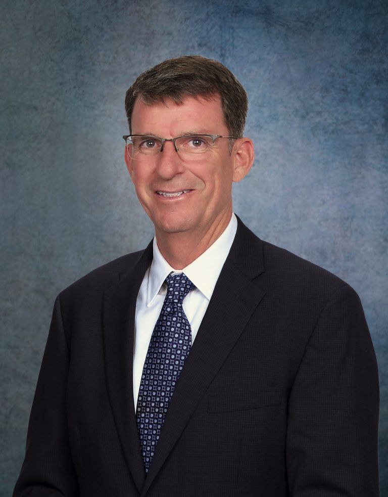 Man in suit and tie, smiling, wearing glasses, against a blurred blue background.