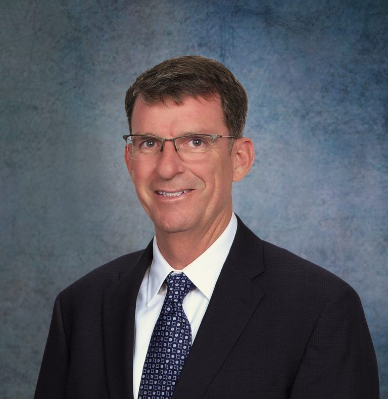 Man in suit and tie, smiling, wearing glasses, against a blurred blue background.