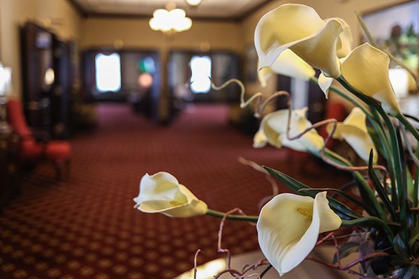 Cream-colored calla lilies in a vase, in front of a long hallway with red patterned carpet and blurred background.