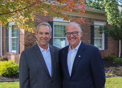 Two men in suits smiling, standing in front of a brick building with fall foliage.