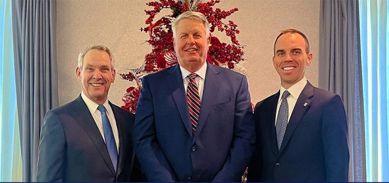 Three men in suits pose smiling in front of a red decorative backdrop.