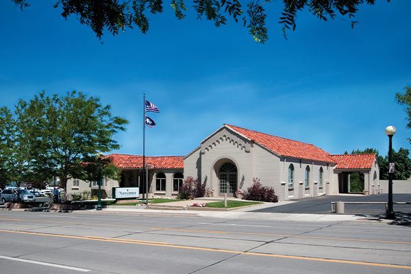 Tan building with red tile roof, two flags. Street view, blue sky, tree.