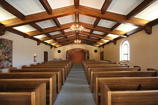 Interior of a chapel with wooden pews and beams, arched doorway, and ornate chandelier.