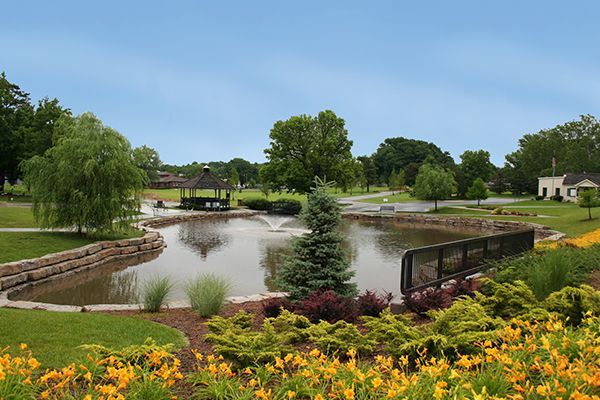 A pond with a gazebo, surrounded by greenery and flowers under a cloudy blue sky.