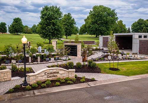Cemetery garden with brick paths, stone walls, and memorial niches. Trees and green grass under a cloudy sky.