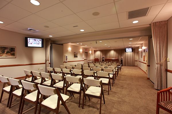 Rows of white chairs face a stage in a beige room. Two small TVs are on the walls.
