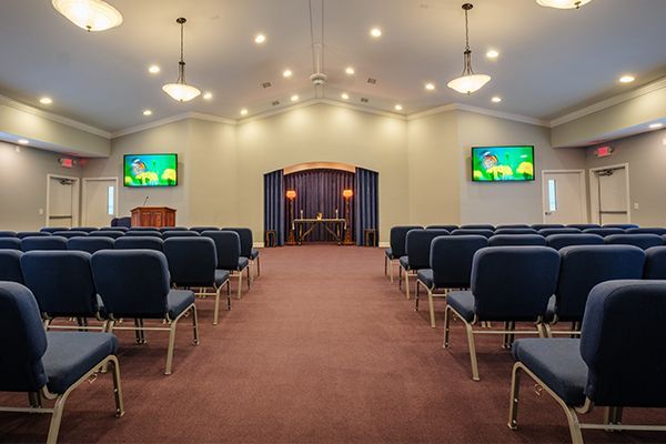 Rows of blue chairs in a chapel face an altar with draped curtains. Two screens display images.