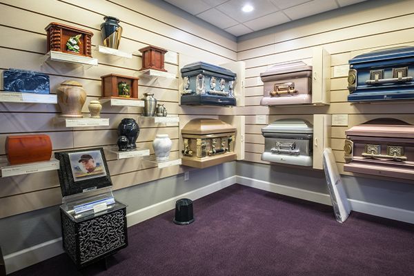 Funeral home showroom displaying caskets and urns on shelves; purple carpet, tan walls.