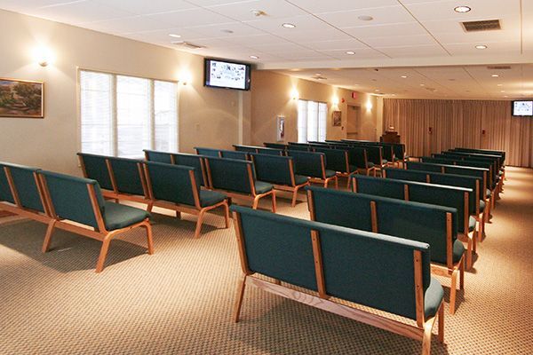 Funeral home chapel with rows of teal seats, beige carpet, and wooden paneling.