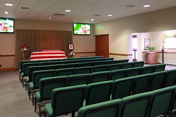 Funeral home interior: rows of green chairs face a casket, two screens display images.