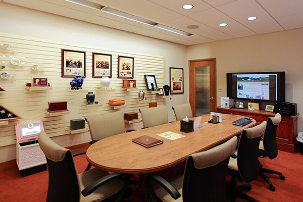 Meeting room with wooden table, chairs, TV, urns and photos on wall.