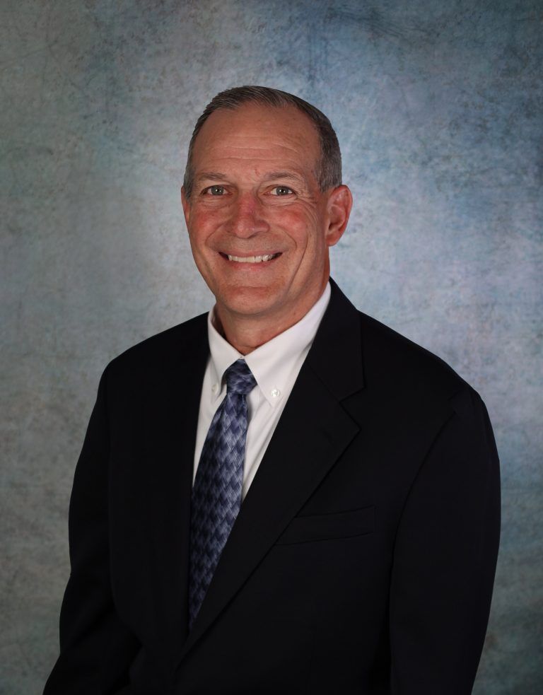 Man in a dark suit and patterned tie smiles, standing against a mottled blue background.